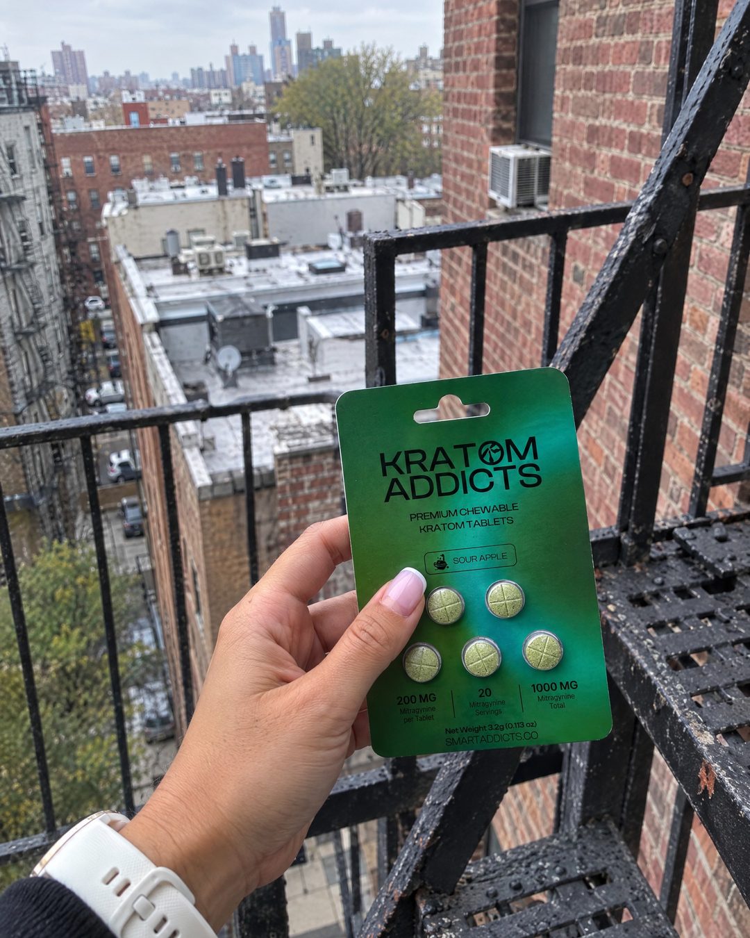 A hand holding Sour Apple kratom tablets on a city fire escape