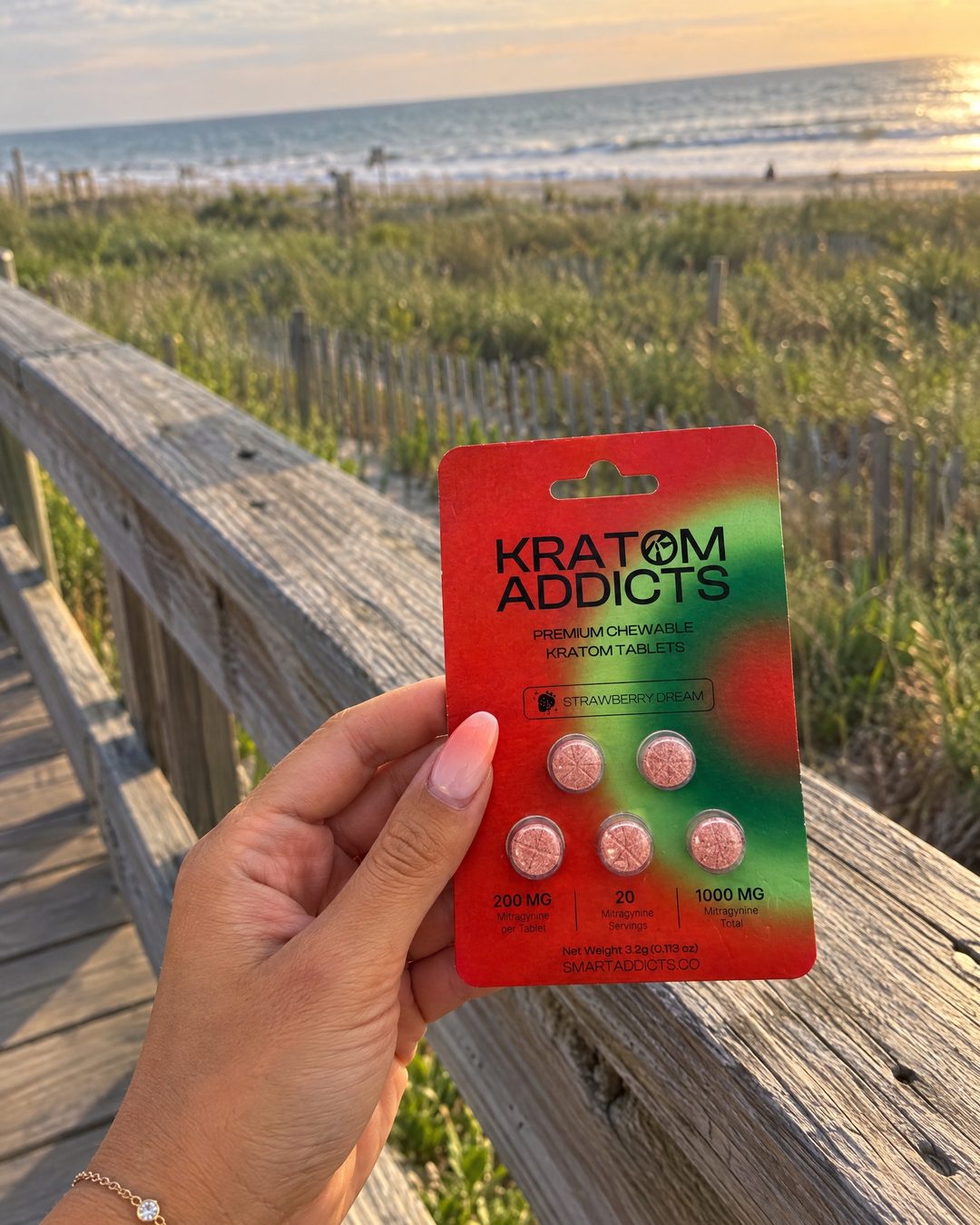A hand holding Strawberry Dream kratom tablets on a boardwalk at sunset