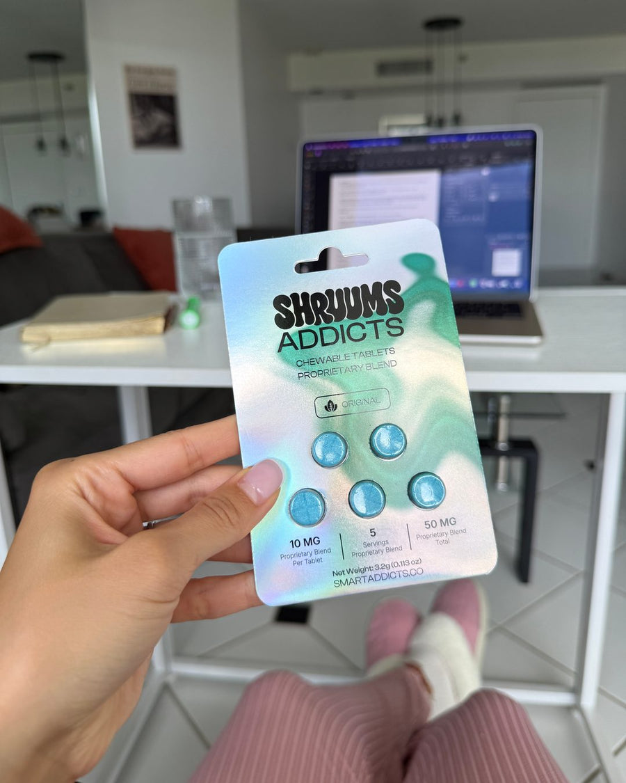 A hand holding Shruums Addicts Original mushroom tablets at a desk, a laptop open behind