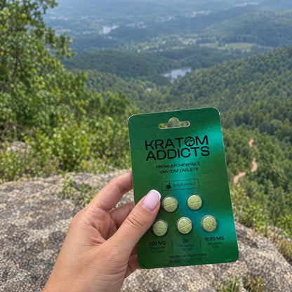 A hand holding Sour Apple kratom tablets above a mountain valley